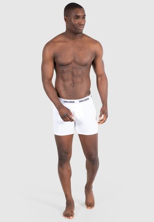 White boxer shorts with a black elastic waistband featuring "SMILODOX" branding. The model stands barefoot, highlighting the shorts' design.