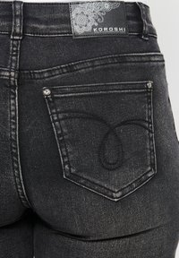 Crne traperice od denima s teksturiranim materijalom, s osebujnim dizajnom na stražnjem džepu i oznakom marke na pojasu.