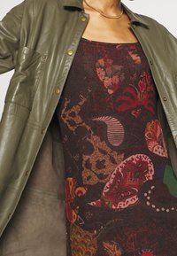 Veste en cuir vert olive avec des boutons-pression, sur une robe sombre à motifs avec des designs floraux et abstraits de couleurs variées.