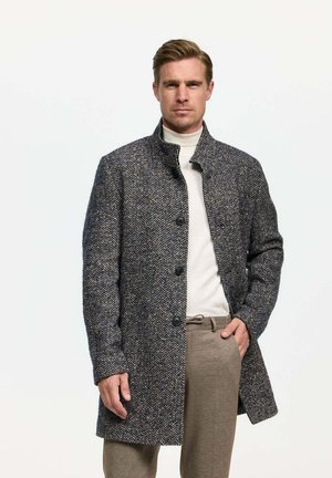 Wollmantel mit Fischgrätmuster in Dunkelblau und Grau, ausgestattet mit einem hohen Kragen, vorderen Knöpfen und einer lässigen Passform. Getragen über einem weißen Pullover.