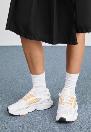 Nogi w białych skarpetkach za kostkę i białych trampkach Adidas z brzoskwiniowymi paskami, stojące na niebieskim dywanie obok czarnego brzegu spódnicy.