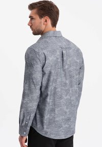 Camisa de patrón gris con diseños florales, de manga larga, con cuello abotonado y detalle de pliegue en la espalda, confeccionada en tejido texturizado.