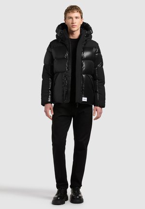 Jeune homme portant une doudoune noire avec capuche, pantalon noir et chaussures noires, debout devant un fond uni gris clair.