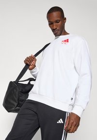 Bílé tričko s červeným grafickým prvkem, spojené s černými kalhotami s bílým logem Adidas. Černá sportovní taška s ramenním popruhem.
