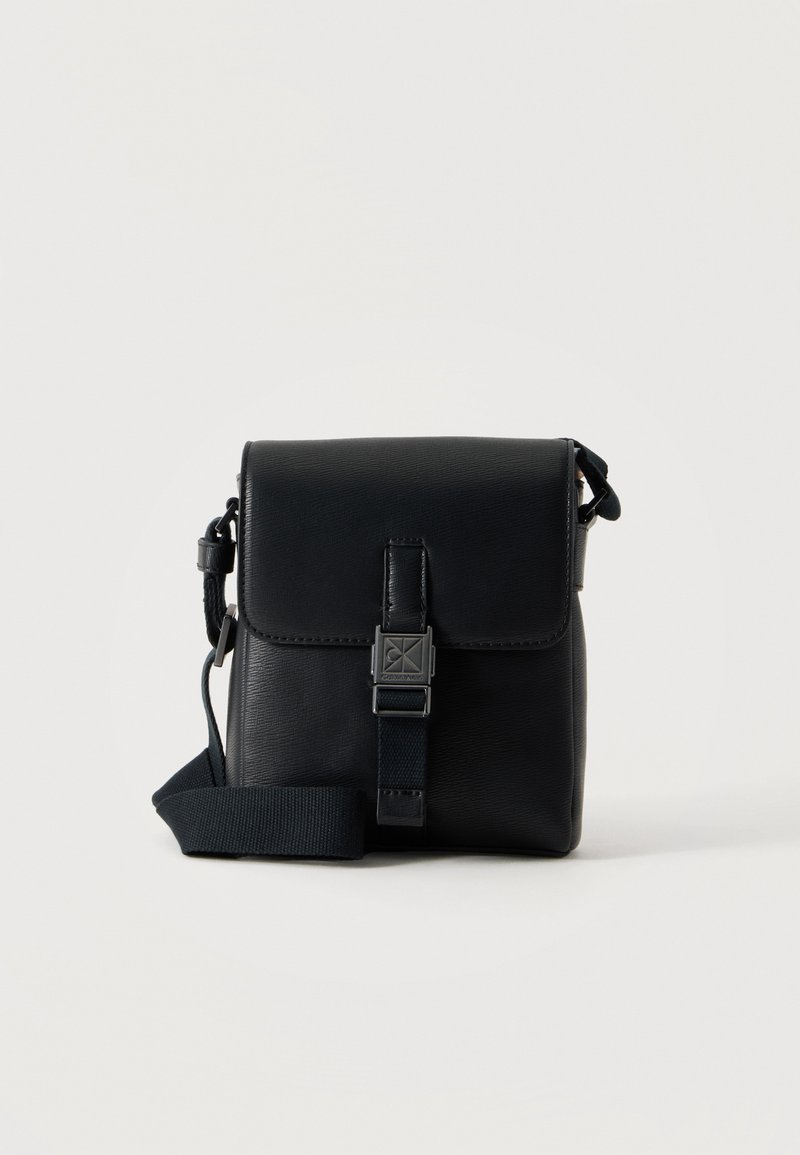 Μικρή μαύρη δερμάτινη τσάντα crossbody με καπάκι και κλείσιμο με αγκράφα, ρυθμιζόμενο λουρί, παρουσιάζεται σε απλό λευκό φόντο.
