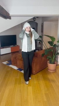 Damespufferjack in lichtgrijs, witte sjaal, zwarte broek en zilveren schoenen, staand in een kamer met een bank en een plant.