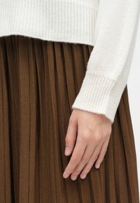 Κρεμ πλεκτό πουλόβερ με ριχτούς καρπούς, συνδυασμένο με καφέ πλισέ φούστα. Εστίαση στην υφή και στις κατακόρυφες γραμμές της φούστας.