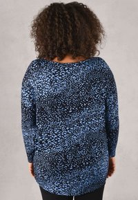 Langarmoberteil aus einem schwarzen Stoff mit blauem Punktmuster. Hat einen weiten Ausschnitt und einen abgerundeten Saum, der eine lockere Passform zeigt.