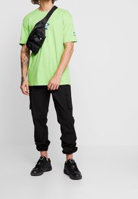 Neon green short-sleeve t-shirt, black cargo pants with pockets, black sneakers, and a black crossbody bag with multiple compartments.