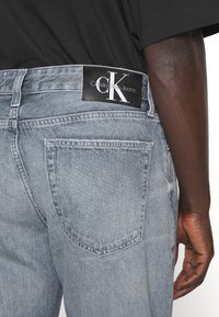 Calvin Klein Jeans, denim azul claro, diseño de pierna recta, cintura de altura media, etiqueta negra en la parte trasera con logo blanco, detalle en el bolsillo trasero.