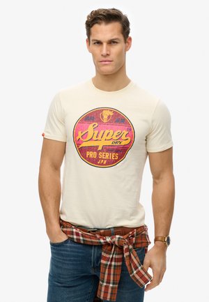 Camiseta crema con un gráfico circular en rojo y amarillo que dice "Super Dry Pro Series". Jeans azules, camisa de cuadros atada a la cintura, reloj casual.