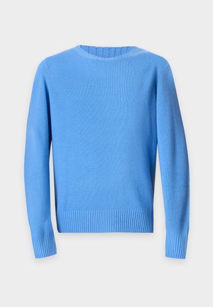 Jeune homme portant un pull bleu, un jean bleu clair avec une ceinture marron et des bottes en cuir foncé, debout devant un fond blanc uni.