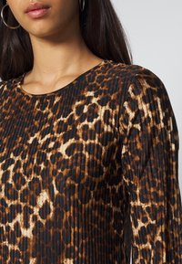 Maglietta a maniche lunghe nera e marrone con design plissettato e motivo a macchie di leopardo, con scollo rotondo. Il tessuto sembra liscio.