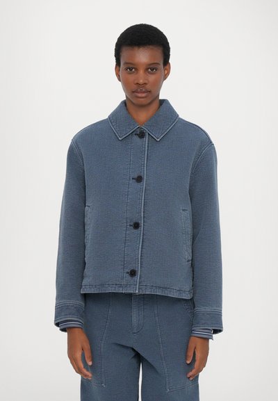 Chaqueta azul con textura, con cuello, botones frontales y dos bolsillos laterales. Presenta un diseño recortado y puños de rayas contrastantes en la parte inferior.
