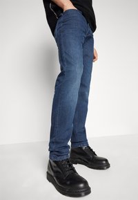 Blaue Denim-Jeans mit schmaler Passform, ausgestattet mit detailliertem Nähen und einer mittelhohen Taille. Kombiniert mit schwarzen Schnürstiefeln.
