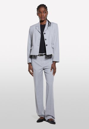 Blazer grigio chiaro su misura con tre bottoni, abbinato a pantaloni a gamba larga coordinati. Top nero sotto, scarpe nere eleganti. Tessuto dalla texture liscia.