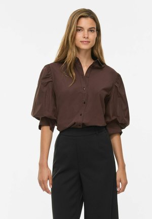 Mujer con cabello largo castaño claro, vestida con una camisa marrón oscuro con mangas abullonadas y pantalones negros de talle alto, de pie sobre un fondo blanco.