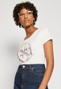 Camiseta blanca de manga corta con un diseño gráfico brillante en la parte frontal, combinada con jeans de mezclilla oscuros de tiro alto. Lleva puestas gafas con montura transparente.