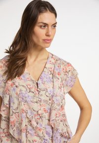 Blusa floreale beige, con motivi viola e rosa. Tessuto morbido, parte frontale arricciata, maniche corte e scollatura a V con dettaglio a bottoni.