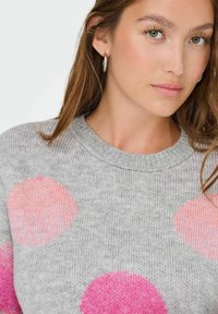 Jeune femme à la peau claire et aux yeux verts portant des boucles d'oreilles en argent et un pull gris avec de grands pois roses et pèches.