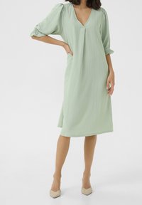 Robe vert clair mi-longue avec un décolleté en V, manches bouffantes, tissu texturé et coupe ample, associée à des escarpins beige à bout pointu.