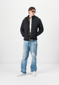 Jachetă neagră, padded, cu glugă și detalii cu capse, purtată cu blugi deschiși la culoare, distresați, și adidași albi, de către o persoană cu ochelari.