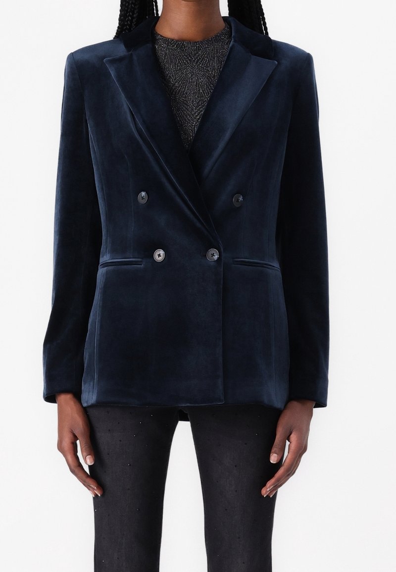 Personne portant un blazer croisé en velours bleu marine à quatre boutons, par-dessus un top noir à motifs et un pantalon noir clouté.
