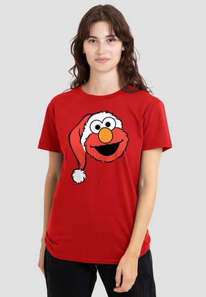 Young woman wearing a red t-shirt with a smiling Elmo character wearing a Santa hat printed on the front, standing against a plain background.