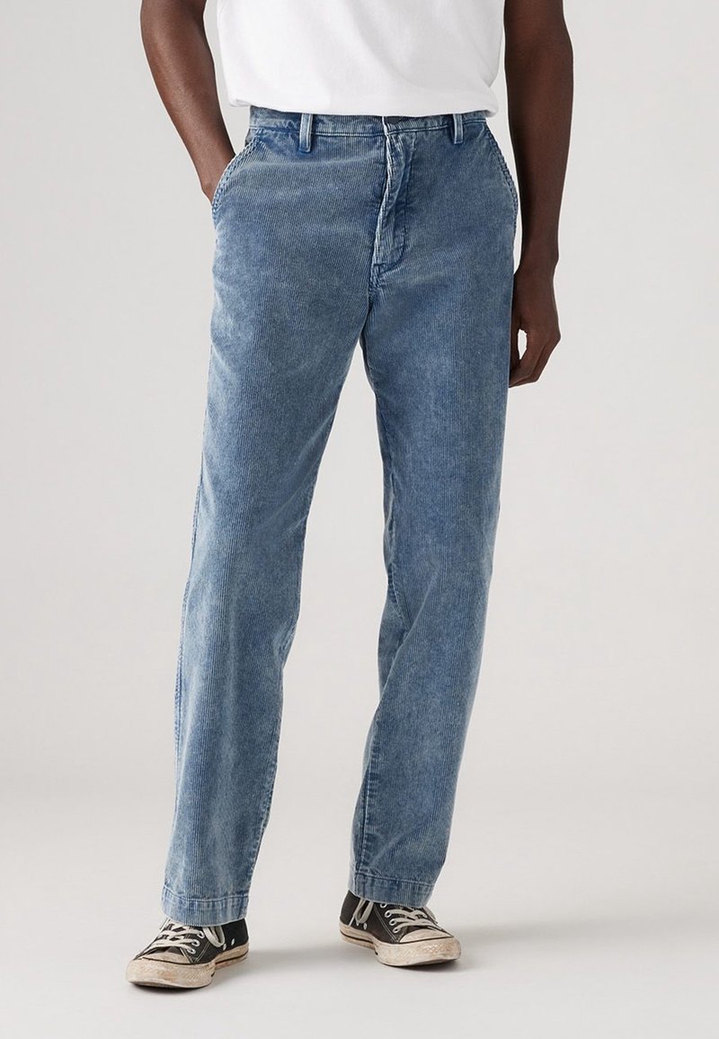 Personne portant un pantalon en velours côtelé bleu clair et des baskets noires à lacets blancs, debout les mains dans les poches devant un fond uni.