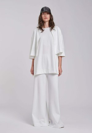 Vrouw draagt een losse witte top met korte mouwen, witte broek met wijde pijpen en een zwarte pet, staand tegen een effen lichte achtergrond.