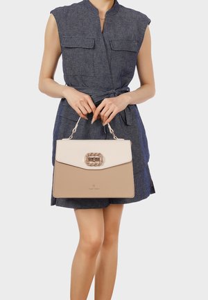 Donna in abito grigio senza maniche che tiene una borsa beige e bianca con chiusura intrecciata davanti a uno sfondo semplice.