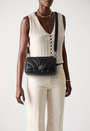 Borsa a tracolla in pelle nera con design trapuntato e dettagli in metallo argentato. Il modello indossa un top in maglia crema e pantaloni avorio.