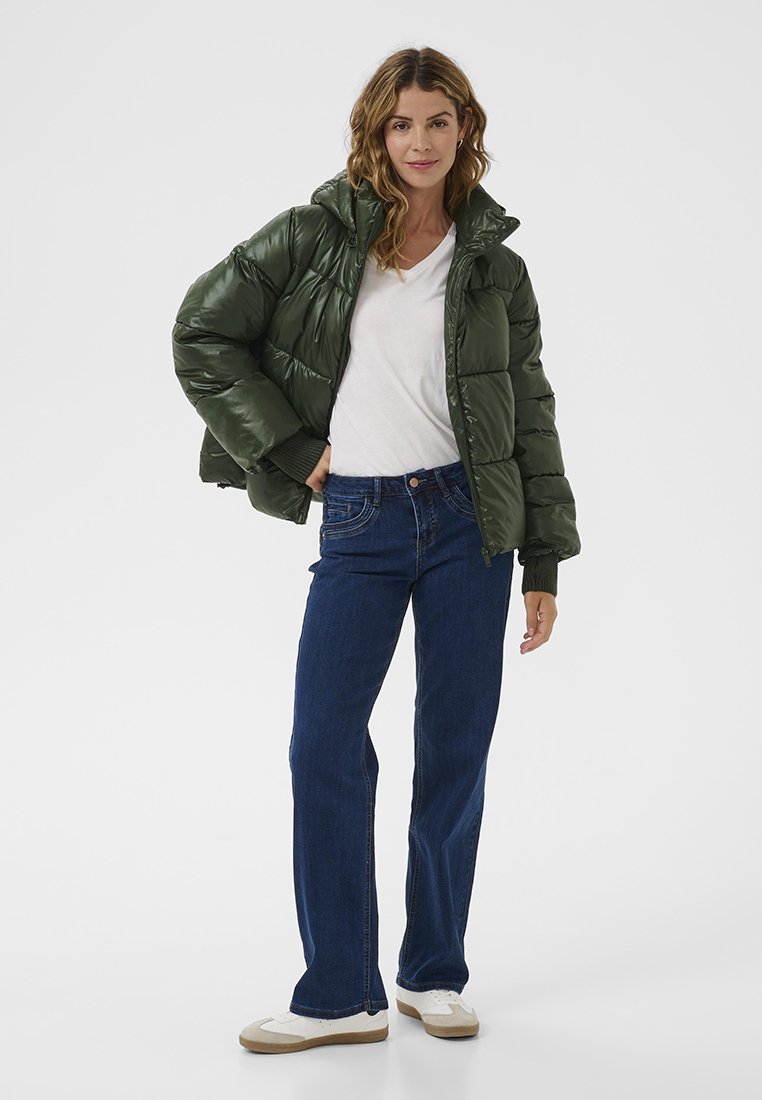 Dunkelgrüne Pufferjacke mit glänzender Oberfläche, ausgestattet mit hohem Kragen und gerippten Manschetten, kombiniert mit blauen Schlagjeans und weißen Sneakers.