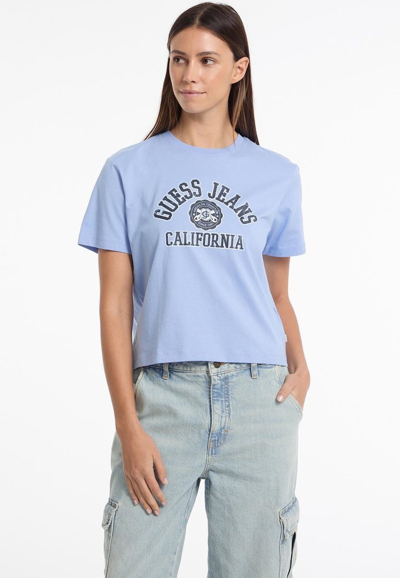 Šviesiai mėlyna trumpa medvilninė marškinėliai su priekiniais spaudiniais, kuriuose yra "Guess Jeans California" tamsiai mėlynomis ir baltomis raidėmis.