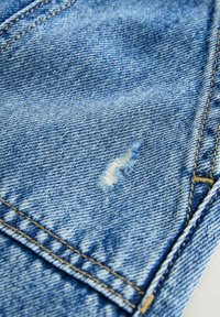 Tejido de denim azul claro con una textura ligeramente desgastada. Presenta un pequeño desgarro cerca del centro, con costuras amarillas a lo largo de las costuras.