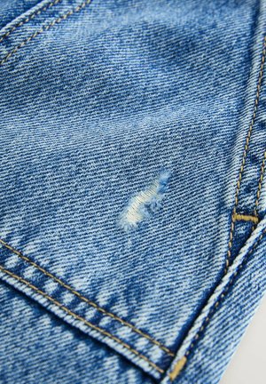 Svetle modrý denimový materiál s mírně vybledlým vzhledem. Obsahuje malou díru poblíž středu a žluté švy podél švů.