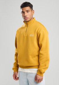 Mustard yellow pullover sweatshirt with a zip neck, ribbed cuffs, and embroidered logo; made from soft, textured cotton fabric.