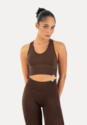 Kobieta z włosami zaczesanymi do tyłu, ubrana w pasujący brązowy bezrękawnik typu crop top i legginsy z wysokim stanem na jednolitym białym tle.