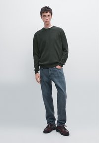 Donkergroene trui met een ronde hals, gecombineerd met lichtblauwe jeans en donkerbruine veterboots. Eenvoudige, casual outfit presentatie.