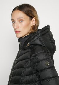 Zwarte donsjack met een hoge kraag en capuchon, voorzien van een horizontaal gestikt patroon, gouden hardware en een logo-accent op de mouw.