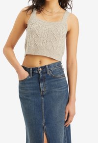 Nainen, jolla on päällään beige neulottu crop top, jossa on tekstuuriset kuviot, sekä korkeavyötäröiset siniset farkkushortsit, joissa on etuhalkio ja käsi taskussa.