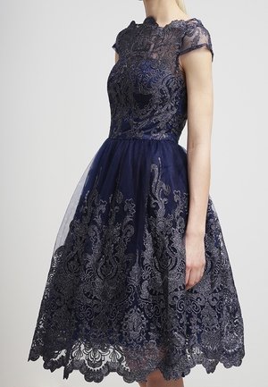 Femme portant une robe en dentelle bleu marine avec des motifs floraux complexes, des manches courtes transparentes et un ourlet festonné.