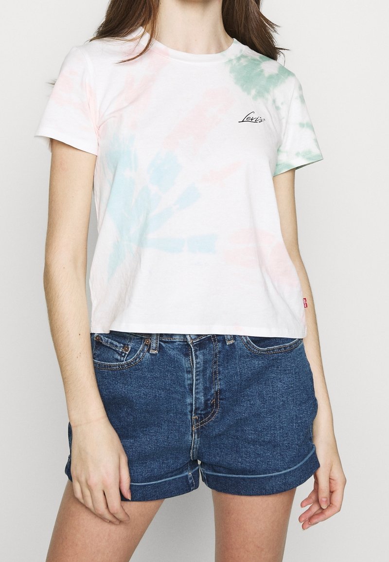 Jeune femme portant un t-shirt blanc tie-dye Levi's et un short en denim bleu, debout les bras détendus le long du corps.