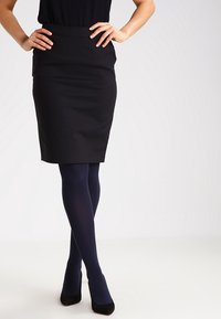 Žena stojící s rukama na bokách, oblečená v černé sukni, tmavě modrých punčochách a černých vysokých podpatcích na jednobarevném pozadí.