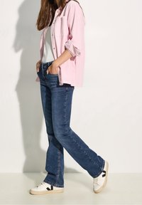 Ljusrosa skjorta med knappar, vit t-shirt, mörkblå jeans och vita sneakers med svarta detaljer. Uppvikta ärmar och avslappnad passform.