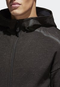 Mörkgrå zip-up hoodie med en texturerad tyg, som har horisontell ribbing och subtila reflektiva detaljer längs axlarna och dragkedjan.