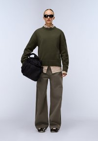 Persona che indossa un maglione oversize verde oliva, una camicia beige, pantaloni a gamba larga, occhiali da sole neri, mentre tiene uno zaino nero su uno sfondo semplice.