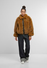 Brūns mākslīgās pūkas mētelis ar augstu apkakli, rāvējslēdzēju un garām piedurknēm. Apvienots ar brīviem tumšiem džinsiem un baltām sporta kurpēm.