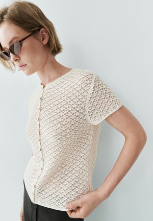 Femme aux cheveux courts portant des lunettes de soleil et un haut blanc texturé à manches courtes boutonné, debout devant un mur clair uni.