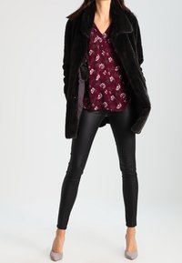 Svart fuskpälsjacka över en burgundy blommig blus, matchat med svarta leggings och ljusgrå spetsiga klackar. Enkla, moderna designelement.